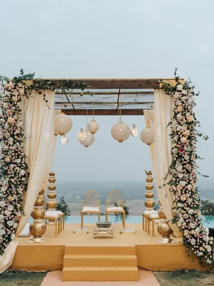 A poolside mandap with a view. The elegant floral pillars and hanging lanterns create a serene and beautiful atmosphere for an evening ceremony.