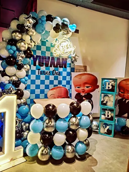 A cool Boss Baby themed first birthday setup. The decoration uses a blue, black, and silver color scheme with a checkered backdrop, a large balloon arch, 'BABY' blocks, and a light-up number '1'.
