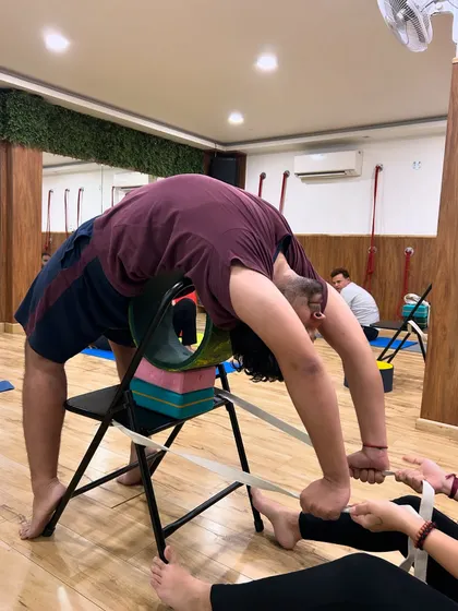 Teamwork in action. A partner provides gentle resistance with a strap while the student uses a chair and wheel to deepen their backbend, ensuring a safe and effective stretch.