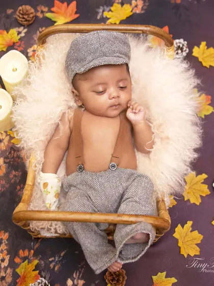 This autumn-themed portrait features a baby boy dressed in a vintage-style flat cap and suspenders. He is resting in a basket surrounded by fall leaves, pinecones, and soft candlelight.