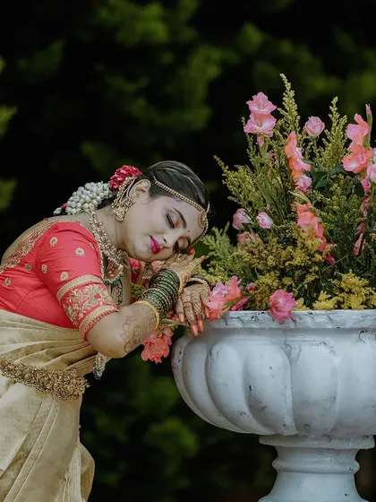 A serene bridal portrait taken outdoors. The bride, dressed in a traditional silk saree, rests against a floral arrangement, creating a peaceful and artistic image that highlights her grace and beauty.