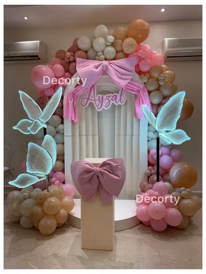 A whimsical butterfly-themed birthday backdrop in shades of pink and peach. The decor features large, glowing neon butterflies, a fabric bow centerpiece, and a beautiful balloon arch.