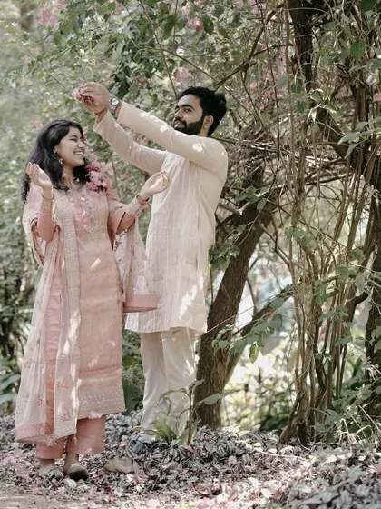A couple enjoys a shower of flower petals in a beautiful garden, both dressed in coordinating pastel traditional outfits.