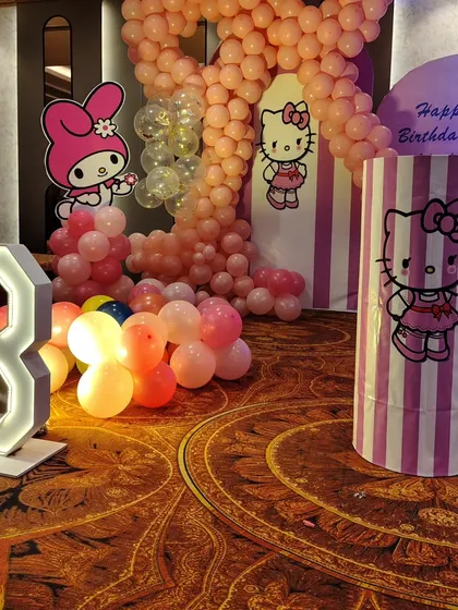 A close-up of the Hello Kitty side of the party stage, with a large balloon number '8', character cutouts, and a pink-and-white striped theme plinth.