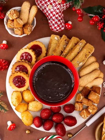 A top-down shot of a Christmas fondue platter, featuring a rich chocolate dip surrounded by cookies, strawberries, and madeleines.