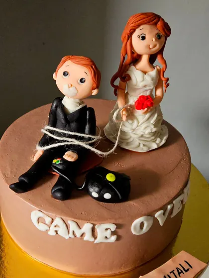 A playful "Game Over" cake for a bachelorette party. It features a humorous scene with handcrafted figurines of the bride and groom, where the groom is tied up with his gaming controller.