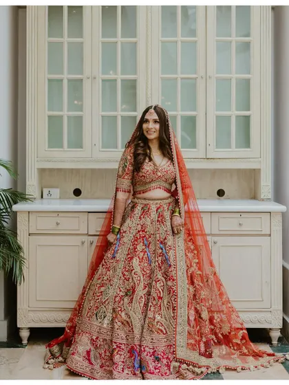 A stunning full-length portrait of a bride in an intricately embroidered red lehenga, standing gracefully in a well-lit room.