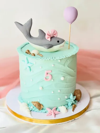 Even sharks can be cute. This "baby shark" inspired cake features a friendly shark with a bow, set in an underwater scene with fondant shells and starfish.