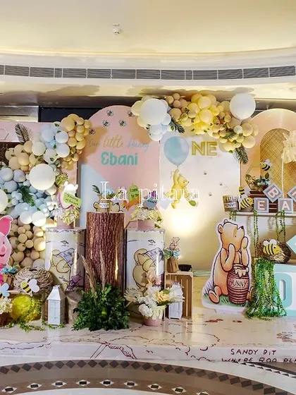 A wide shot of a charming Winnie the Pooh first birthday party. The decor features a soft, pastel color palette, a backdrop of the Hundred Acre Wood, and adorable cutouts of Pooh, Piglet, and their friends.