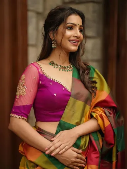 A model pairs a multi-colored saree with a magenta blouse featuring sheer, embroidered sleeves, complemented by a green beaded necklace.