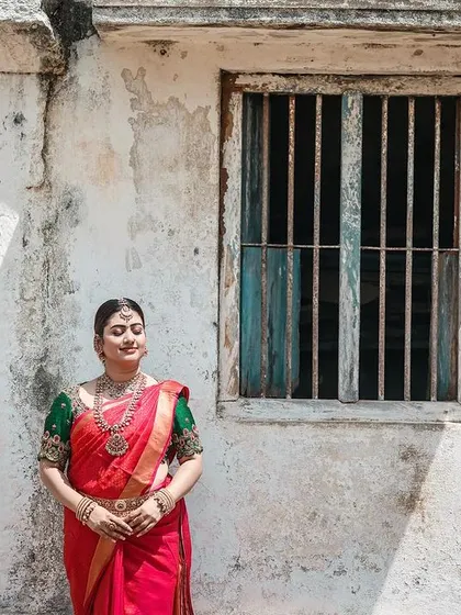 An environmental portrait of a model in a red saree against a weathered, rustic building. This shot combines traditional fashion with an interesting, textured background.
