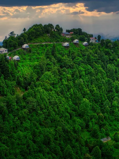 A wide shot of the Kanatal dome stay, capturing the property under dramatic sun rays breaking through the clouds. This image highlights the magical, dream-like quality of a glamping adventure.