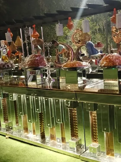 An expansive buffet setup on a lush green lawn for an outdoor event. The long, mirrored counter reflects the ambient light, creating a beautiful effect in the evening.