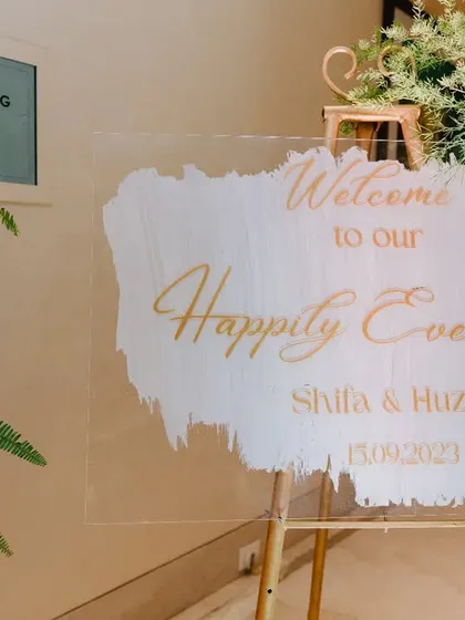A personalized acrylic welcome sign for Shifa and Huzaifa's "Happily Ever After," elegantly scripted in gold and surrounded by lush floral arrangements.