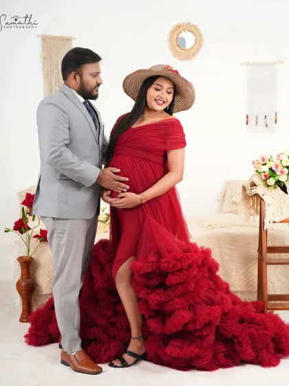 A stylish couple portrait with the mother-to-be in a red gown and hat. The boho-chic studio setting provides a trendy and romantic backdrop.