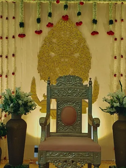An elegant and regal seating arrangement for a traditional ceremony. A single ornate chair is placed against a backdrop of jasmine garlands and a golden carved panel, creating a simple yet powerful focal point.