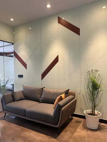A comfortable grey sofa in a waiting area, set against a back-painted glass wall with abstract brown line details. A potted palm adds a touch of nature to this contemporary corner.