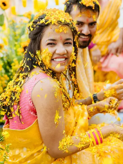 A radiant portrait of the bride, her face covered in turmeric and flowers, smiling brightly for the camera.