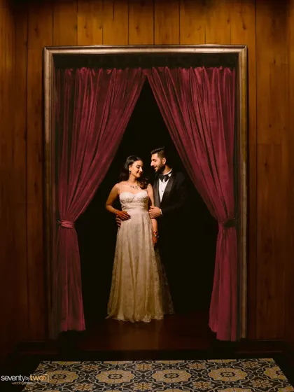 A dramatic portrait of the couple framed by rich, red curtains, kicking off their wedding celebrations with an evening of elegance in Siliguri.