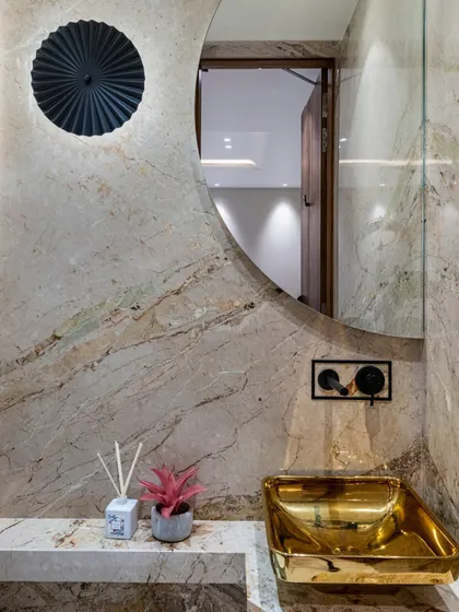 A powder room that exudes glamour. A metallic gold sink is set against a natural stone wall, with a circular mirror and a modern black light fixture adding to the drama.