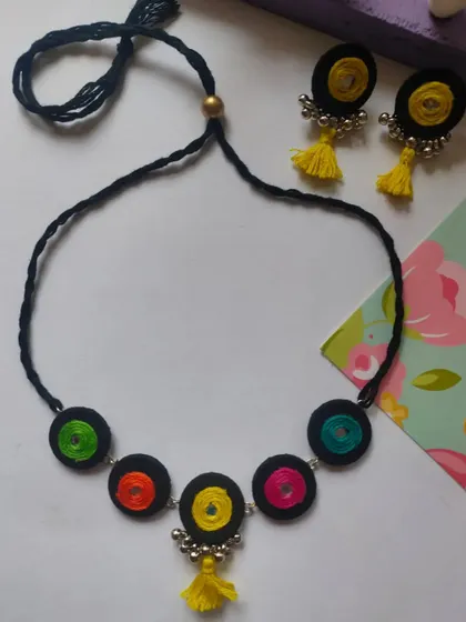 A playful and colorful handmade necklace set. This design features thread-wrapped circles in multiple colors on a black cord, with matching earrings that have bright yellow tassels.