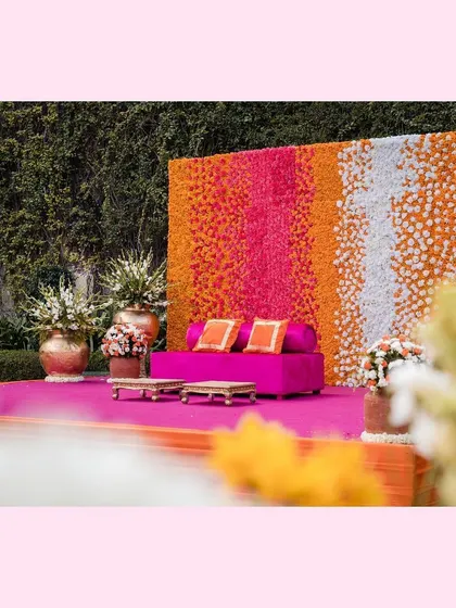 A vibrant Mehendi stage with a backdrop of colorful flowers and a bright pink sofa.