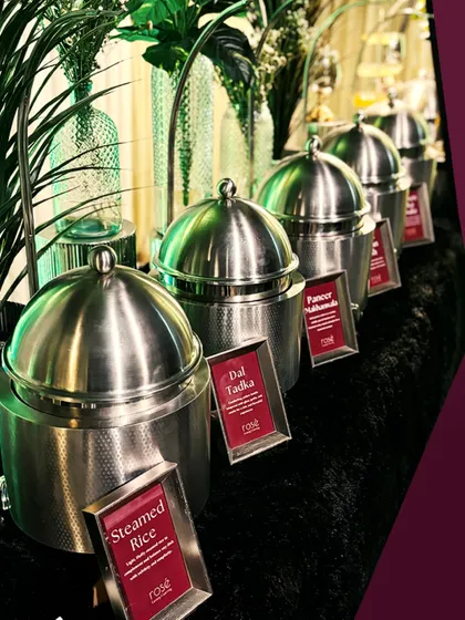 A line of modern, stainless steel chafing dishes holding our Indian main courses like Steamed Rice and Dal Tadka. The clean lines and lush green backdrop provide a contemporary and fresh look for your event.