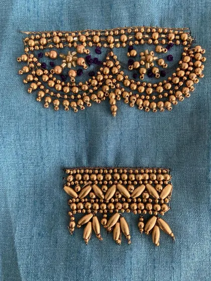 Two distinct embroidery patterns on blue fabric, showcasing different beading and threadwork techniques. The top design is a classic neckline pattern, while the bottom is a border style.