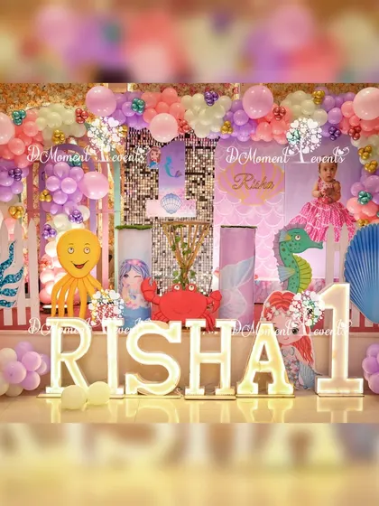 A full and elaborate mermaid-themed stage for a first birthday, featuring a sequin backdrop, light-up letters, and a variety of cute sea animal props like an octopus and crab.