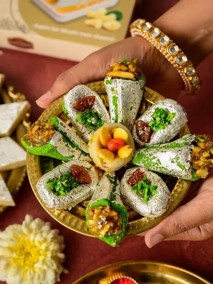A festive platter of Indian sweets, captured with rich, warm lighting to evoke a sense of tradition and celebration. The detailed styling with edible silver leaf and pistachios highlights the craftsmanship of the product.