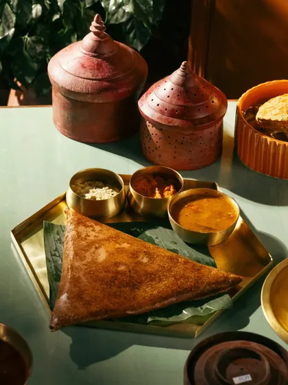 A crispy ghee roast dosa with sambar and chutney. The moody lighting and traditional props create a rich, atmospheric scene.
