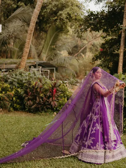 A dramatic shot of a bride in a purple lehenga, her long, flowing veil spread out behind her in a lush green garden setting.