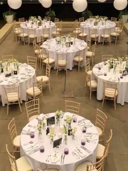 A simple and elegant guest dining setup for a terrace event. The round tables are covered in white linen, with classic Chiavari chairs and minimalist floral centerpieces.