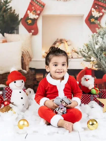 A toddler girl in a Santa suit smiles happily in a festive Christmas setting.