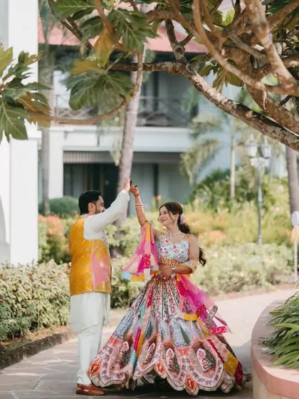 A couple shares a dance under a tree, the groom twirling the bride in her vibrant, multi-colored lehenga during their Haldi celebration.