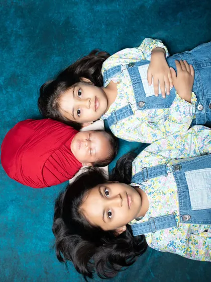 An overhead shot of two sisters lying next to their newborn sibling, who is wrapped snugly in a red swaddle.