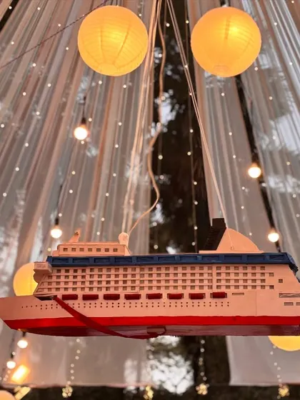 A unique decor element for a travel-themed or nautical-themed wedding. This model cruise ship hanging amidst lanterns adds a personalized and whimsical touch to the celebration.