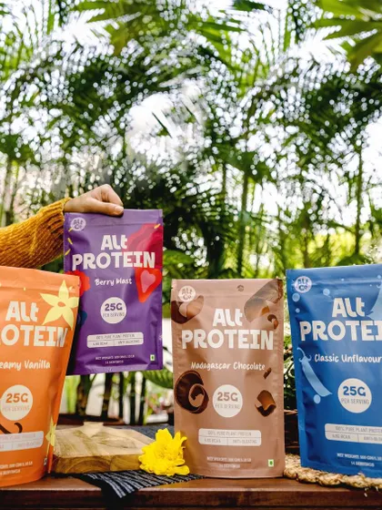 A colorful lineup of Alt Co.'s different protein powder flavors. The outdoor, natural setting and the hand holding up the packets create a friendly and approachable feel.