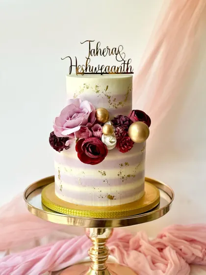 A beautiful two-tier cake with purple and white striped buttercream and gold leaf accents. It's decorated with a mix of silk flowers and gold spheres for a modern, celebratory look.