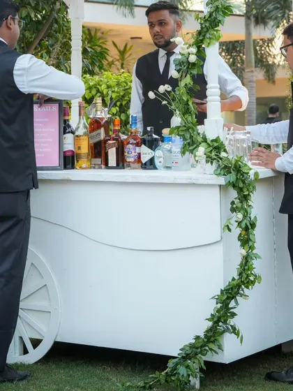 A charming white bar cart adorned with a simple garland of greenery and white roses, ready to serve celebratory drinks at the sundowner event.