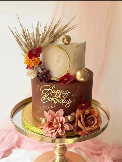 A stylish two-tier birthday cake combining a rich chocolate base and a marbled white top tier. The cake is decorated with dried pampas grass, silk flowers, and gold accents for a modern bohemian feel.