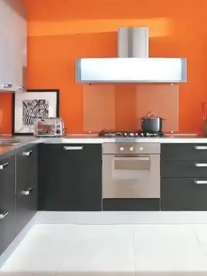 A bold L-shaped kitchen with a vibrant orange accent wall. The dark base cabinets and stainless steel appliances create a strong, contemporary look.