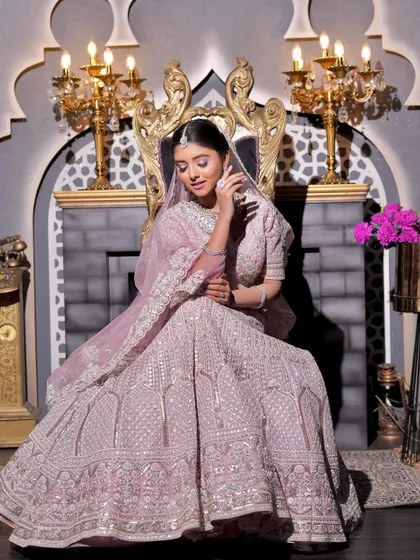 A majestic shot of the bride seated on a throne-like chair. The full view of her lilac-pink lehenga and the grand setting is complemented by her flawless, regal makeup.