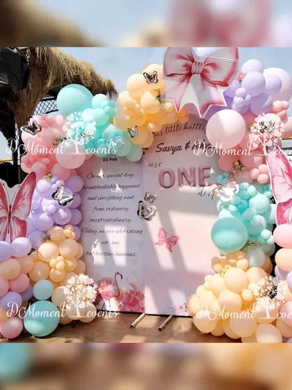 A lovely butterfly-themed backdrop for a twins' first birthday, using soft pastel balloons and large butterfly wing props to frame a personalized message board.