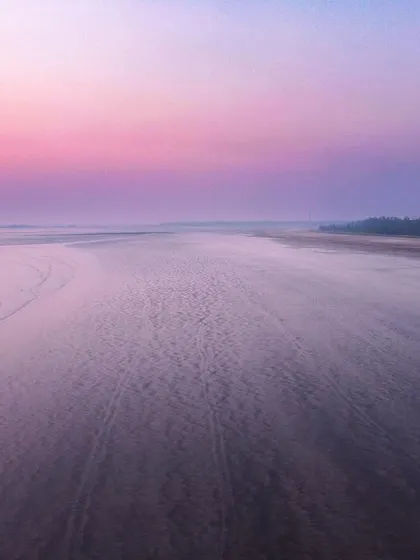 A wide, empty expanse of sand and shallow water is bathed in a purple and pink glow, evoking the feeling of a beach on another planet.