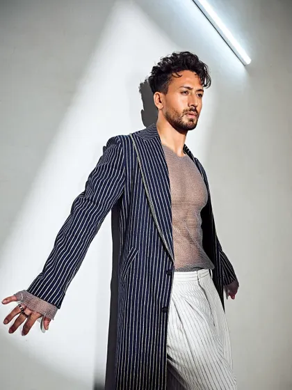 A high-fashion, edgy look for Tiger Shroff. The combination of the long pinstripe coat, sheer top, and white trousers is bold and unexpected.