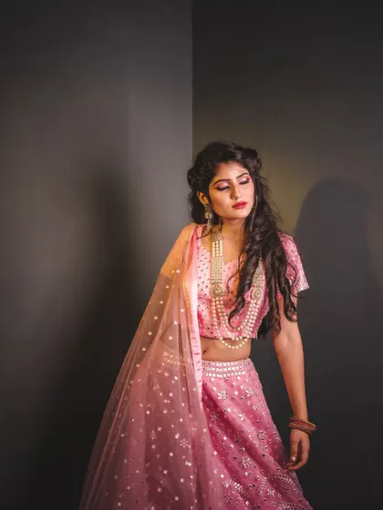 A contemplative pose from a fashion shoot. The soft lighting and the model's expression create a quiet, elegant mood, highlighting the delicate pearl necklace and the texture of the pink lehenga.