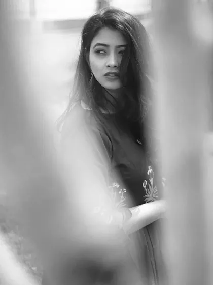 Peeking through the blurred foreground of tree trunks, this black and white portrait creates a sense of a private, stolen moment. The model's sideways glance and the natural framing give the image a mysterious and artistic quality.