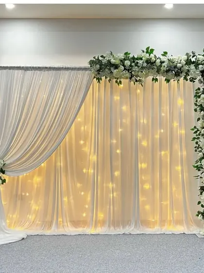 A beautiful and clean backdrop for a baby shower or naming ceremony. This setup uses white drapes with fairy lights and is framed by a corner arrangement of white florals and greenery.