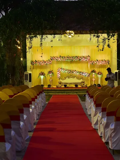 A wide view of an outdoor reception setup at night. The stage is beautifully lit, and the red carpet aisle with neat rows of chairs creates a formal and inviting space for the celebration.